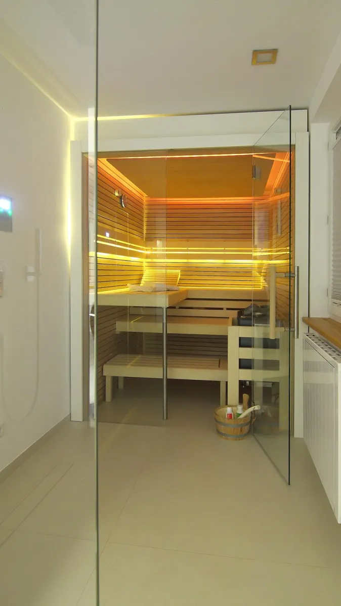 Helles Badezimmer mit integrierter Sauna hinter einer Glastür, moderner Beleuchtung und klassischem Saunaaufguss-Eimer im Vordergrund.