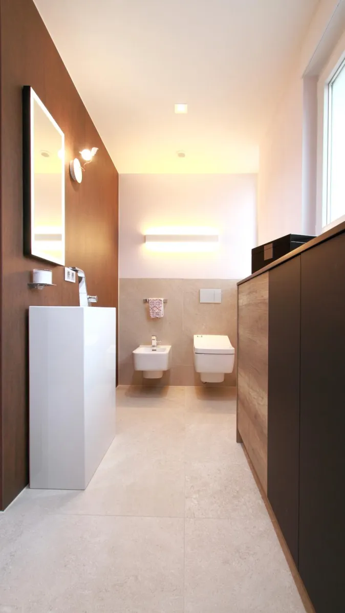 Modernes Wohnbad mit Bidet und WC, wandhängend montiert, kombiniert mit schlichtem Waschtisch, Holzverkleidung und eleganter Beleuchtung.