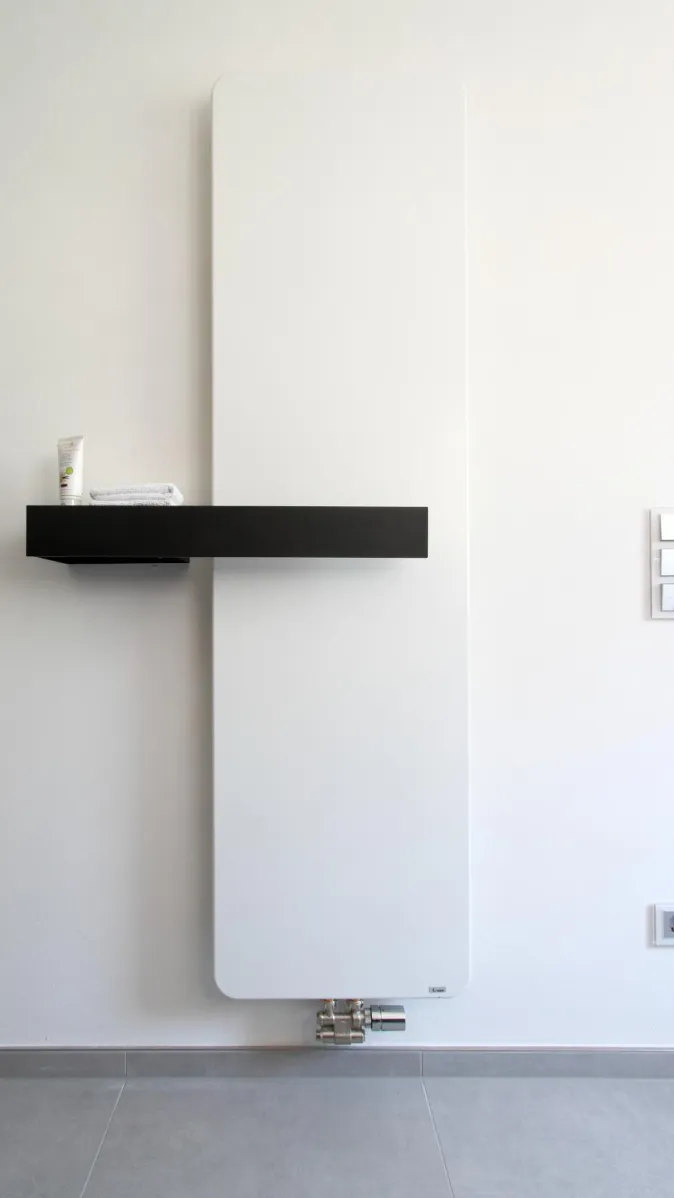 Moderner Designheizkörper in Weiß mit integrierter schwarzer Ablage, stilvoll installiert in einem Bad in Betonoptik.