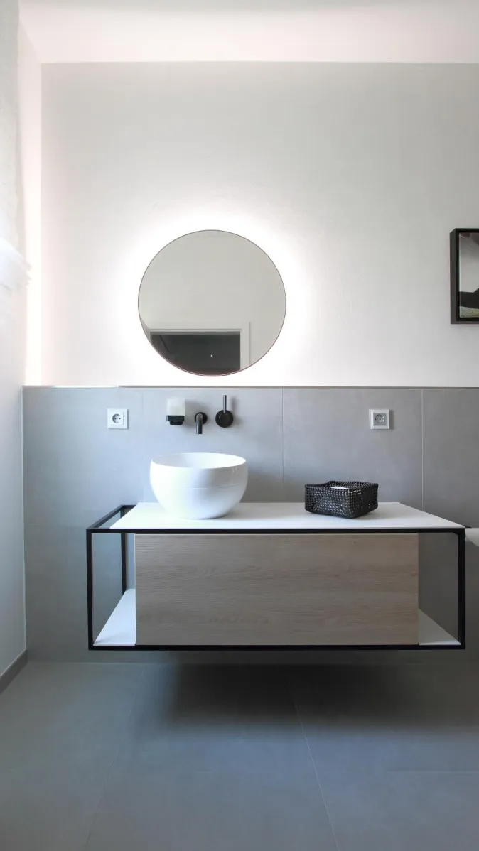 Modernes Badezimmer in Betonoptik mit rundem LED-Spiegel, minimalistischem Aufsatzwaschbecken und schwebendem Waschtisch mit Holzfront.