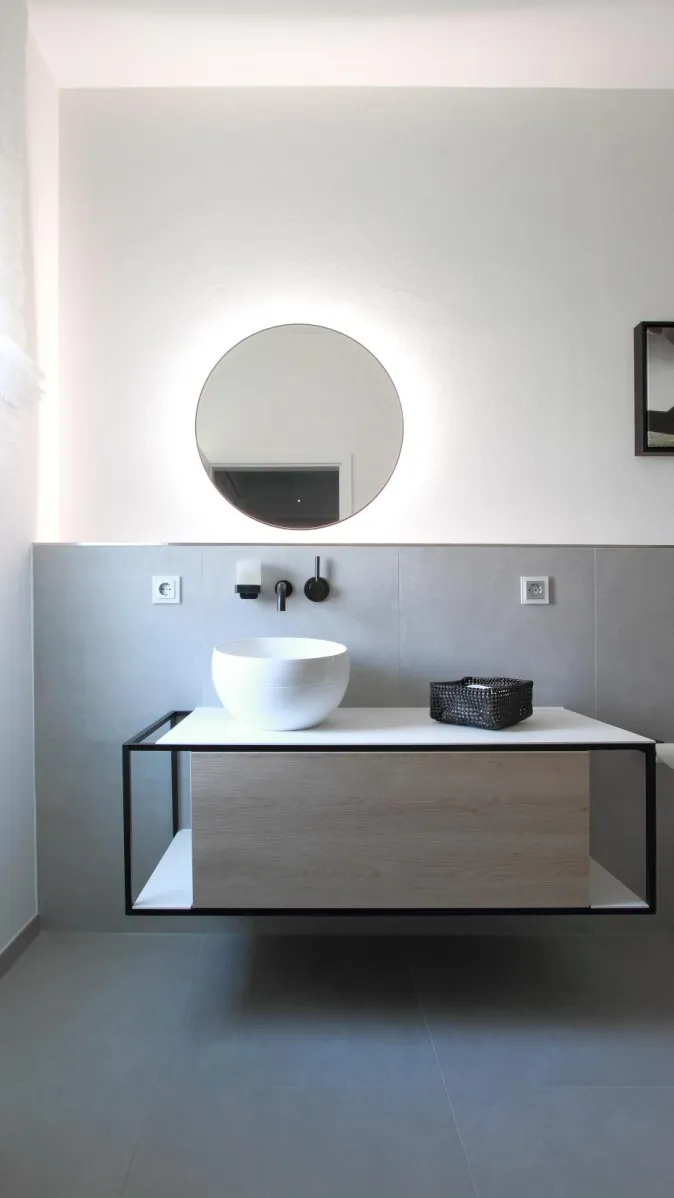 Modernes Badezimmer in Betonoptik mit rundem LED-Spiegel, minimalistischem Aufsatzwaschbecken und schwebendem Waschtisch mit Holzfront.