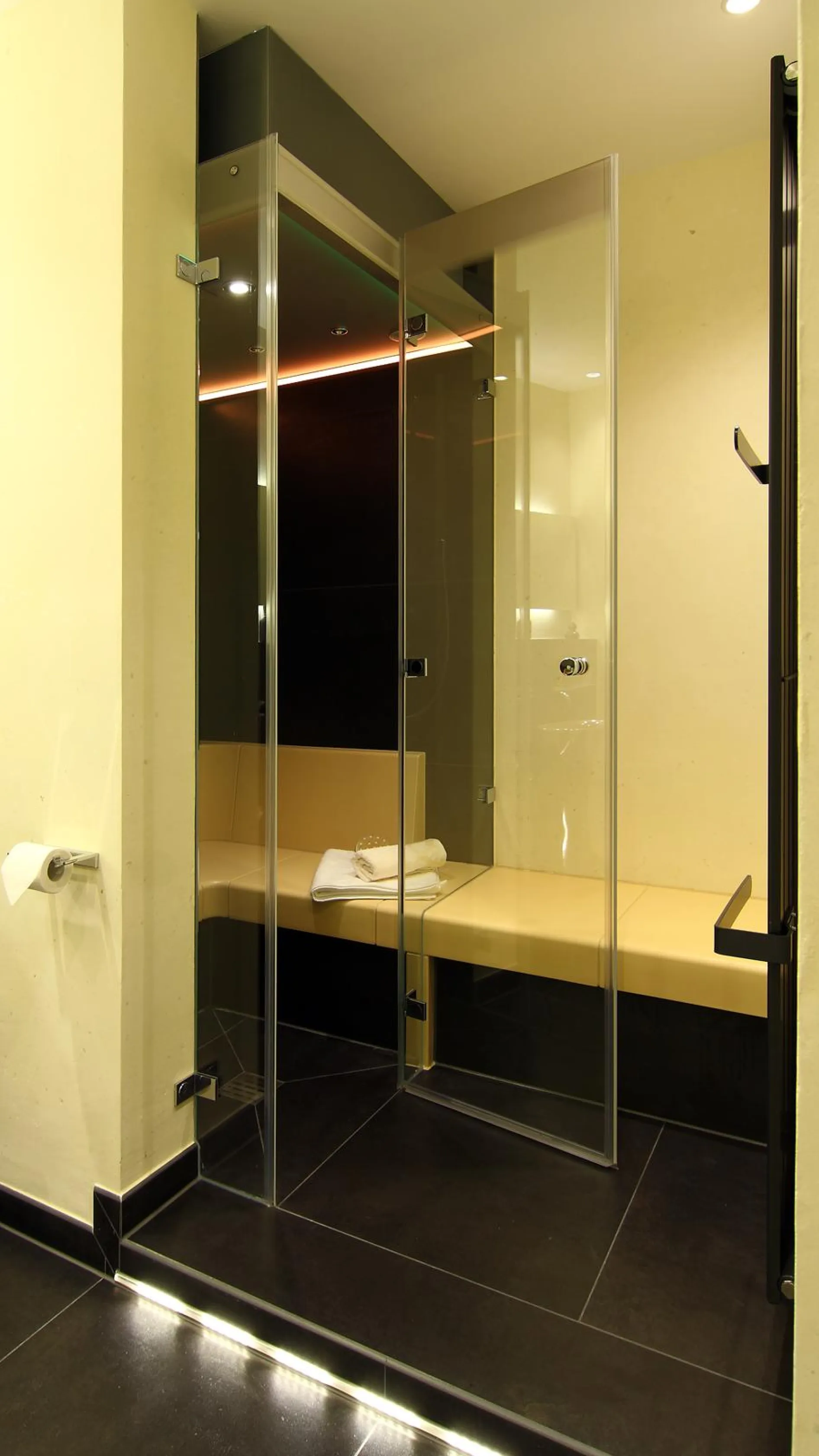 Edles Dampfbad mit heller Sitzbank, Glasfront und integrierter Dusche, modern beleuchtet und mit Handtuch auf der Bank.