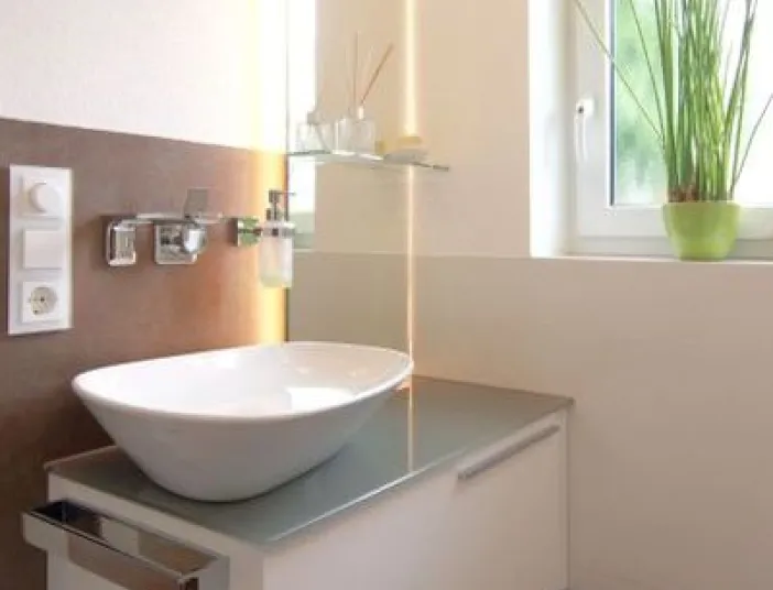 Helles Gäste-WC mit modernem Aufsatzwaschbecken auf weißem Unterschrank, wandmontierter Armatur und dekorativen Details am Fenster.
