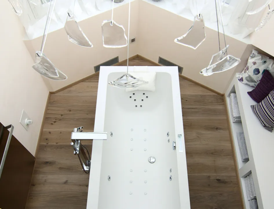 Vogelperspektive auf eine moderne Badewanne mit Whirlpool-Funktion, Holzfußboden, dekorativen Kissen auf der Fensterbank und stilvoller Beleuchtung.