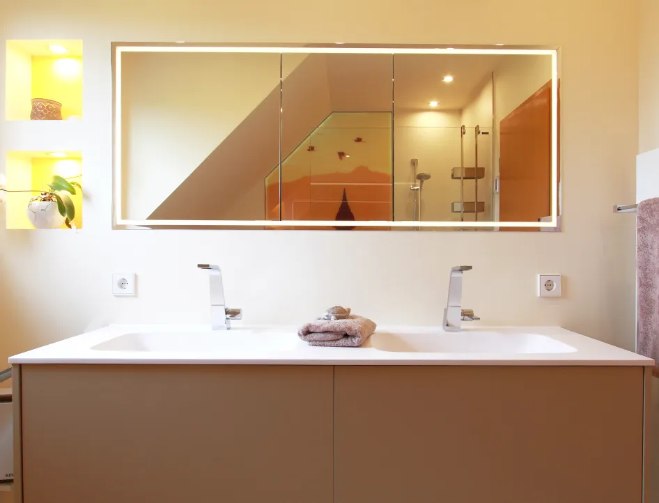 Moderner Doppelwaschtisch mit zwei Chromarmaturen, großem beleuchtetem Spiegelschrank und Blick auf die bodenebene Dusche mit Wandmotiv im Wohnbad.