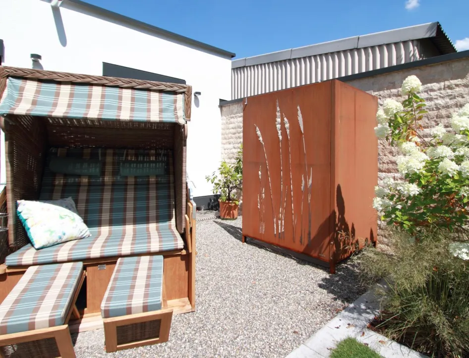 Außenbereich eines Wellnessbads mit Strandkorb, dekorativer Sichtschutzwand und blühenden Pflanzen als Rückzugsort nach der Sauna.