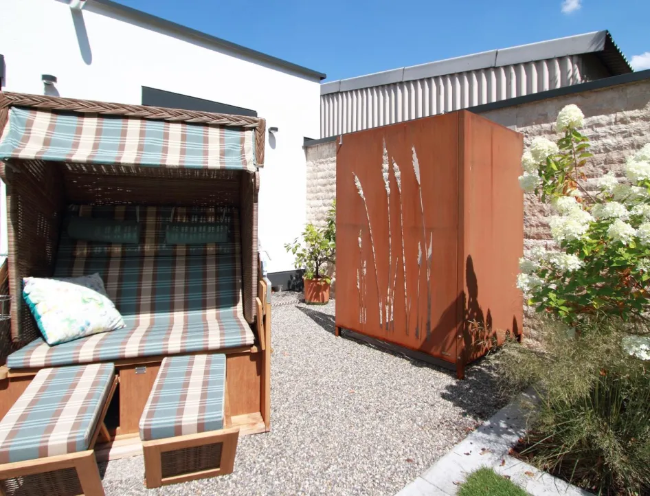 Außenbereich eines Wellnessbads mit Strandkorb, dekorativer Sichtschutzwand und blühenden Pflanzen als Rückzugsort nach der Sauna.