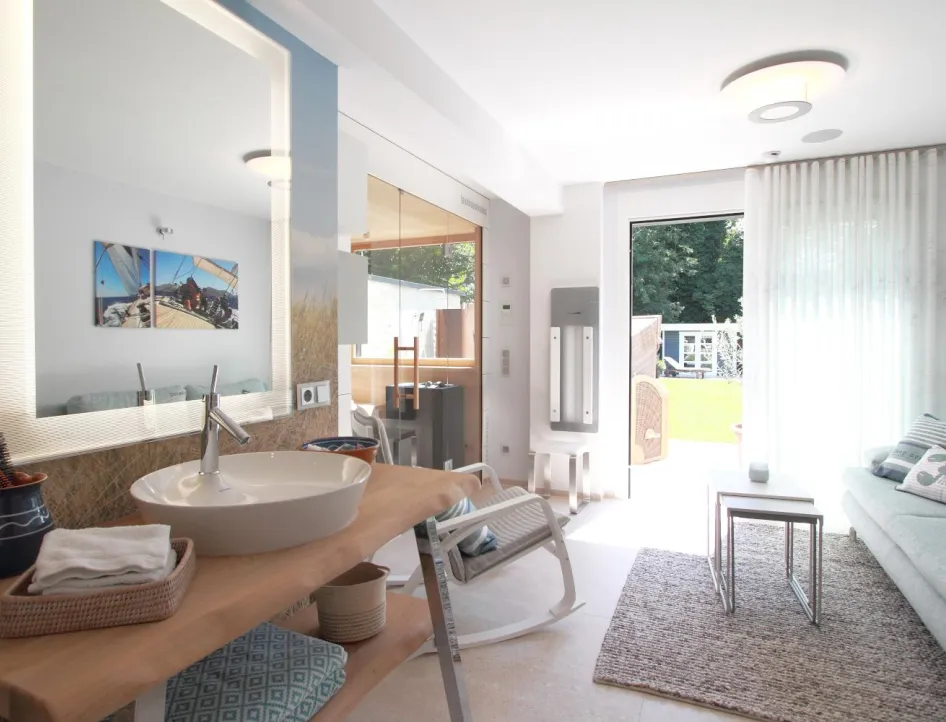 Helles Wellnessbad mit beleuchtetem Spiegel, offenem Waschtisch, gemütlicher Sitzecke und angrenzender Sauna mit Blick ins Grüne.
