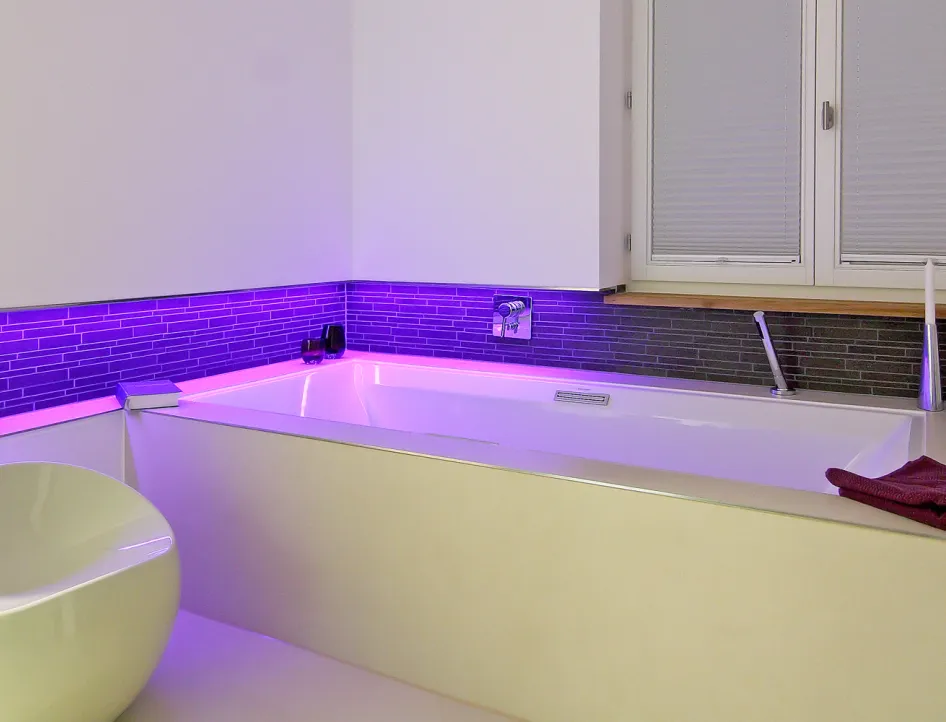 Badewanne mit moderner LED-Beleuchtung in Lila, grauer Fliesenrückwand und stilvoller Einrichtung in einem Wellnessbad.